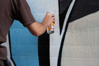 A person is spray painting a design on a wall, focusing on precise lines and shapes in shades of blue, black, and white. The image captures the lower arm of the person holding the spray can.