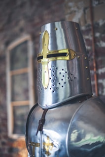 A close-up of a medieval knight's helmet and armor, featuring a metallic design with gold accents and intricate detailing. The background is softly blurred, suggesting an interior setting with a textured wall.