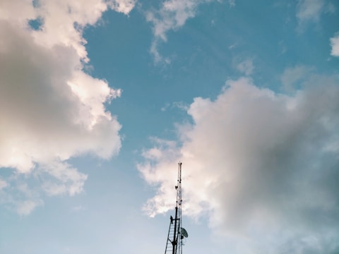 A tall communication tower stands against a backdrop of a bright blue sky with scattered, fluffy white and gray clouds. The structure is centered in the image, reaching up into the sky.