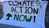 A large white banner displays the bold text 'Climate Action Now!' in black and green. A green tree silhouette is painted alongside the words. The banner appears to be mounted on a metal structure, suggesting it is part of a protest or public demonstration.