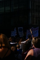 A group of people gathered outside a building, holding signs with messages about science and unity. The scene suggests a protest or rally, with diverse individuals participating. Some signs are partially visible, displaying phrases like 'Science Says' and 'One World'. The setting suggests a city environment with glass windows reflecting light.