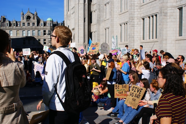 A large group of people is gathered outside a historic building, participating in a protest. Many are holding signs with messages related to climate change, advocating for environmental action, and expressing concern for the future. The crowd includes people of various ages, and they appear engaged in the demonstration. The building has a classic architectural style with ornate features.