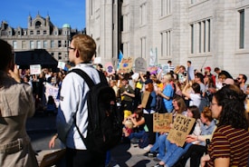 A large group of people is gathered outside a historic building, participating in a protest. Many are holding signs with messages related to climate change, advocating for environmental action, and expressing concern for the future. The crowd includes people of various ages, and they appear engaged in the demonstration. The building has a classic architectural style with ornate features.
