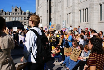 A large group of people is gathered outside a historic building, participating in a protest. Many are holding signs with messages related to climate change, advocating for environmental action, and expressing concern for the future. The crowd includes people of various ages, and they appear engaged in the demonstration. The building has a classic architectural style with ornate features.