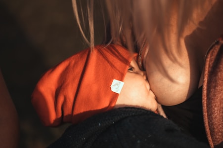 A baby in a red hat is breastfeeding, captured in a close-up shot. The lighting is warm, highlighting the baby and the caregiver's skin tone.
