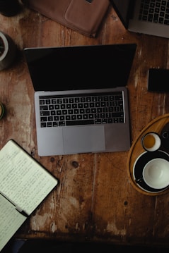 A wooden table holds a laptop with a closed screen, an open notebook filled with handwriting, a leather pouch, and a tray with empty and partially filled cups. The setting suggests a workspace with a rustic and cozy ambiance.