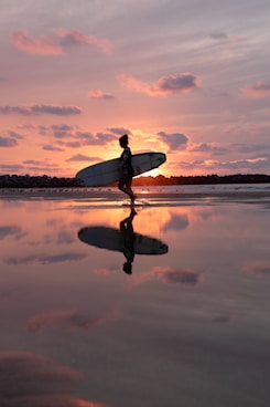 reflection of walking woman holding surfboard on body of water during golden hour