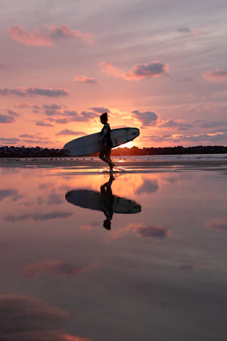 reflection of walking woman holding surfboard on body of water during golden hour