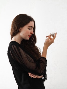 A woman with long wavy hair is holding a small perfume bottle in her left hand while looking at it intently. She is wearing a black blouse with sheer, dotted sleeves and has a pensive expression on her face.