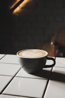 A cup of coffee with latte art sits on a white tiled surface. The cup is dark, contrasting with the creamy beige pattern on top of the coffee. The background is dimly lit, adding a cozy and intimate atmosphere.