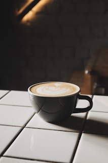 A cup of coffee with latte art sits on a white tiled surface. The cup is dark, contrasting with the creamy beige pattern on top of the coffee. The background is dimly lit, adding a cozy and intimate atmosphere.
