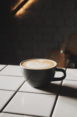 A cup of coffee with latte art sits on a white tiled surface. The cup is dark, contrasting with the creamy beige pattern on top of the coffee. The background is dimly lit, adding a cozy and intimate atmosphere.