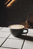 A cup of coffee with latte art sits on a white tiled surface. The cup is dark, contrasting with the creamy beige pattern on top of the coffee. The background is dimly lit, adding a cozy and intimate atmosphere.