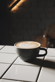A cup of coffee with latte art sits on a white tiled surface. The cup is dark, contrasting with the creamy beige pattern on top of the coffee. The background is dimly lit, adding a cozy and intimate atmosphere.