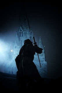 A silhouette of a statue holding a sword is set against the backdrop of a tall, gothic-style church tower. The scene is enveloped in a misty blue ambiance, creating a mysterious and dramatic atmosphere.
