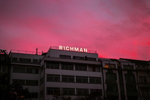 A building with multiple stories features the word 'RICHMAN' in illuminated letters on its rooftop. The sky above is a blend of vibrant pink and purple hues, suggesting either sunrise or sunset, casting a dramatic backdrop.