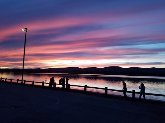 A serene lakeside setting during sunset, featuring vibrant pink, purple, and blue hues in the sky reflected upon the water. Silhouetted figures are present near the shore, possibly taking photographs or enjoying the view. A lamppost is prominently visible in the foreground, contrasting with the natural landscape and the distant hills.
