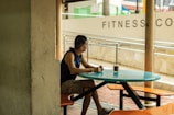A person is sitting at a turquoise table in an outdoor setting, looking off to the side. The surroundings include a brick walkway, a cream-colored wall with the words FITNESS visible, and some poles. The person is wearing a sleeveless shirt and shorts, with one arm resting on the table, next to a glass of beverage.