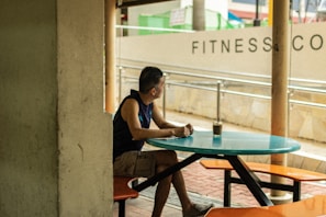 A person is sitting at a turquoise table in an outdoor setting, looking off to the side. The surroundings include a brick walkway, a cream-colored wall with the words FITNESS visible, and some poles. The person is wearing a sleeveless shirt and shorts, with one arm resting on the table, next to a glass of beverage.