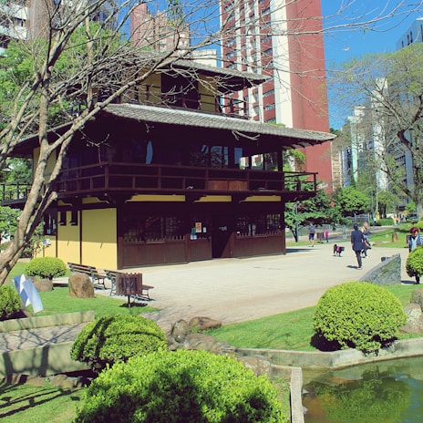 A traditional Japanese-style building surrounded by a garden with neatly trimmed bushes and trees. People are walking along a path, and a small pond is visible in the foreground. Modern high-rise buildings are in the background under a clear blue sky.