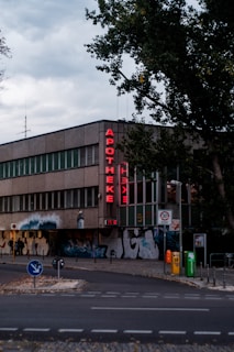 A building with a neon sign reading 'APOTHEKE' is visible on the street corner. The building exterior features modern architecture with glass and stone elements. A row of windows is aligned on the upper floor, with street art covering part of the ground level. A tree partially obscures the view on the right. Traffic signs, a few trash bins in various colors, and a speed limit sign are present on the pavement. The sky is overcast, giving the scene a subdued lighting.