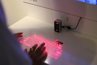 A person is interacting with a virtual keyboard projected onto a flat surface. The keyboard is illuminated with red light from a small device on the desk, which is plugged into an outlet. The hands are positioned as if typing on the projected keyboard.