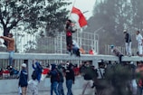 A group of people are engaged in a protest or demonstration. Some individuals are wearing masks and holding flags, while others appear to be shouting or raising their fists. They are gathered near a fence with trees in the background, and a few are climbing on the structure.
