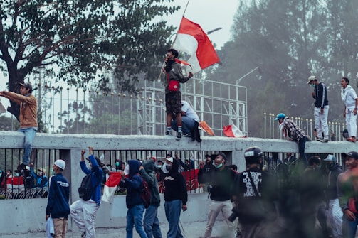 A group of people are engaged in a protest or demonstration. Some individuals are wearing masks and holding flags, while others appear to be shouting or raising their fists. They are gathered near a fence with trees in the background, and a few are climbing on the structure.