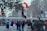 A group of people are engaged in a protest or demonstration. Some individuals are wearing masks and holding flags, while others appear to be shouting or raising their fists. They are gathered near a fence with trees in the background, and a few are climbing on the structure.