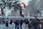 A group of people are engaged in a protest or demonstration. Some individuals are wearing masks and holding flags, while others appear to be shouting or raising their fists. They are gathered near a fence with trees in the background, and a few are climbing on the structure.