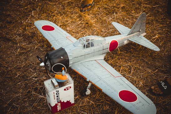 A model airplane with Japanese insignia is resting on the ground beside a fuel container. The plane features a light blue-gray body with red circular markings. It is set on dry, straw-covered ground and there is a partial view of a person’s foot in sandals nearby.