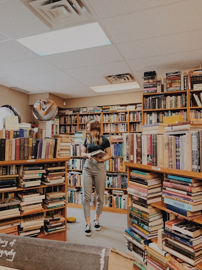 A person stands in a cozy bookstore surrounded by shelves filled with a diverse selection of books. The individual holds a book, appearing to be engaged in reading or browsing. The room is warmly lit with overhead lights, and the ambiance feels inviting and peaceful. The floor is carpeted, and a variety of book genres can be seen around the space.