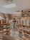 A person stands in a cozy bookstore surrounded by shelves filled with a diverse selection of books. The individual holds a book, appearing to be engaged in reading or browsing. The room is warmly lit with overhead lights, and the ambiance feels inviting and peaceful. The floor is carpeted, and a variety of book genres can be seen around the space.