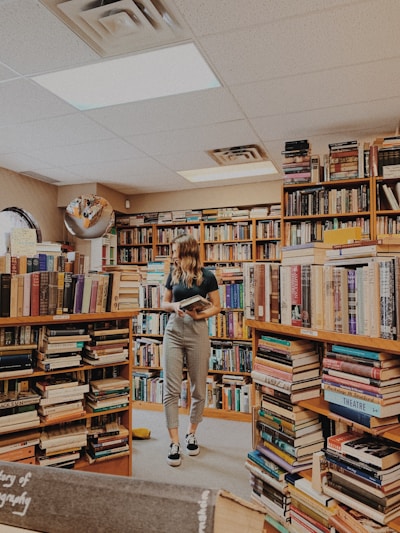 A person stands in a cozy bookstore surrounded by shelves filled with a diverse selection of books. The individual holds a book, appearing to be engaged in reading or browsing. The room is warmly lit with overhead lights, and the ambiance feels inviting and peaceful. The floor is carpeted, and a variety of book genres can be seen around the space.