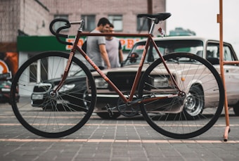A classic-style bicycle with a slender frame is positioned in the foreground on a paved area, with a handrail visible on the right side. In the background, two individuals stand near parked vintage and modern cars, against the backdrop of a brick building with a storefront sign.