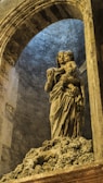 A stone sculpture of a woman holding a child, situated within an arched architectural setting. The sculpture has intricate details in the drapery and facial expressions, conveying a sense of protection and serenity. The stone surface is weathered, adding texture and depth to the image. The setting has a mysterious ambiance, illuminated by subtle lighting that enhances the solemnity of the scene.