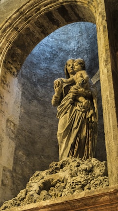 A stone sculpture of a woman holding a child, situated within an arched architectural setting. The sculpture has intricate details in the drapery and facial expressions, conveying a sense of protection and serenity. The stone surface is weathered, adding texture and depth to the image. The setting has a mysterious ambiance, illuminated by subtle lighting that enhances the solemnity of the scene.