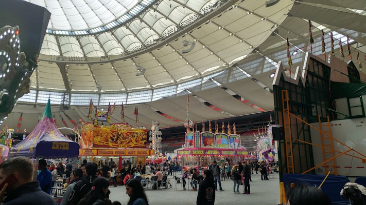 A bustling indoor carnival scene featuring food stalls and colorful attractions under a large, domed roof. There are signs advertising gourmet fries, hot dogs, and various other snacks. The area is filled with people milling about, enjoying the festivities. Bright lights and vibrant decorations create a lively atmosphere.