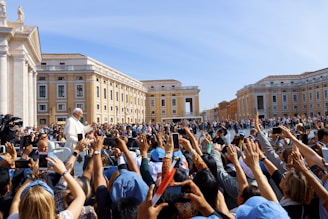 A large crowd is gathered around a prominent religious figure dressed in white, likely a leader or a figure of authority. The people in the crowd are holding up phones and cameras to take photos. The surrounding architecture is grand and historic, suggesting a significant location.