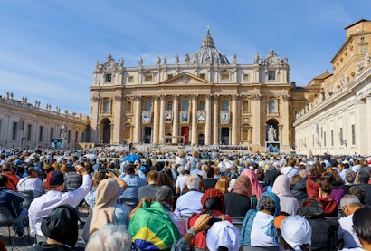 A large crowd gathers in front of an ornate, historic building with columns and statues. The building prominently displays several large portraits. The crowd is diverse, with people wearing various types of clothing including headscarves and hats.