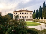 A grand, historic villa with classical architecture is situated amidst a lush garden. The building has an ornate, symmetrical facade featuring statues and decorative elements. A pathway encircles a green lawn leading to the entrance. Tall trees and blooming flowers enhance the scenic backdrop.