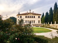 A grand, historic villa with classical architecture is situated amidst a lush garden. The building has an ornate, symmetrical facade featuring statues and decorative elements. A pathway encircles a green lawn leading to the entrance. Tall trees and blooming flowers enhance the scenic backdrop.