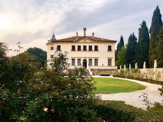 A grand, historic villa with classical architecture is situated amidst a lush garden. The building has an ornate, symmetrical facade featuring statues and decorative elements. A pathway encircles a green lawn leading to the entrance. Tall trees and blooming flowers enhance the scenic backdrop.