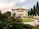 A grand, historic villa with classical architecture is situated amidst a lush garden. The building has an ornate, symmetrical facade featuring statues and decorative elements. A pathway encircles a green lawn leading to the entrance. Tall trees and blooming flowers enhance the scenic backdrop.