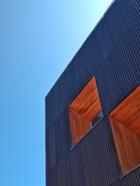 A modern building facade featuring vertical dark metal slats and large, recessed wooden sections. The clear blue sky provides a contrasting backdrop.