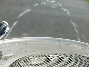Water arcs elegantly from a polished drinking fountain onto a metallic grate. The fountain nozzle is shiny, reflecting the light, while the water forms a smooth arc before descending to the drain below. The background is blurred, focusing attention on the water and fountain.
