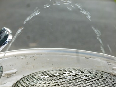 Water arcs elegantly from a polished drinking fountain onto a metallic grate. The fountain nozzle is shiny, reflecting the light, while the water forms a smooth arc before descending to the drain below. The background is blurred, focusing attention on the water and fountain.