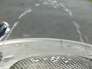 Water arcs elegantly from a polished drinking fountain onto a metallic grate. The fountain nozzle is shiny, reflecting the light, while the water forms a smooth arc before descending to the drain below. The background is blurred, focusing attention on the water and fountain.