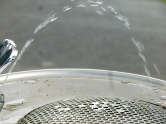 Water arcs elegantly from a polished drinking fountain onto a metallic grate. The fountain nozzle is shiny, reflecting the light, while the water forms a smooth arc before descending to the drain below. The background is blurred, focusing attention on the water and fountain.