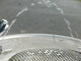 Water arcs elegantly from a polished drinking fountain onto a metallic grate. The fountain nozzle is shiny, reflecting the light, while the water forms a smooth arc before descending to the drain below. The background is blurred, focusing attention on the water and fountain.